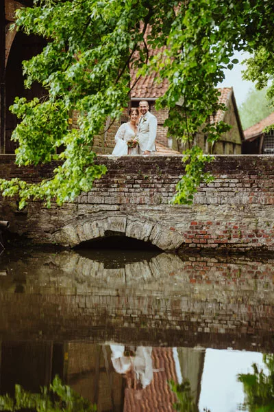 Brautpaar auf einer alten Steinbrücke hinter grünen Blättern mit Spiegelung im Wasser.