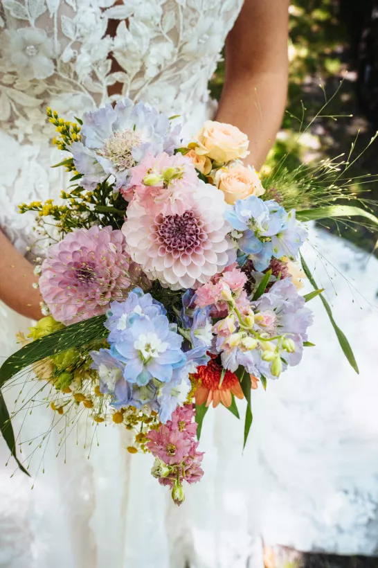 Bunter Brautstrauß mit rosa Dahlien und blauen Blüten vor weißem Spitzenkleid