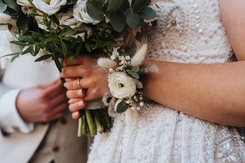 Detailaufnahme der Hände einer Braut mit weißem Blumenstrauß und floralem Armschmuck vor einem Spitzenkleid