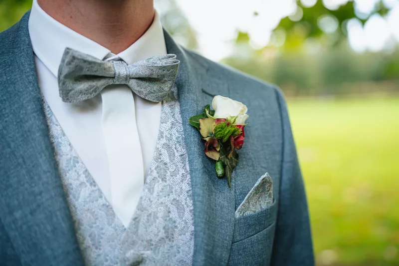 Detailaufnahme eines hellblauen Anzugs mit einer weißen Rosen-Ansteckblume und grauer Fliege.