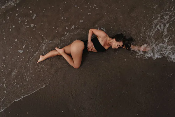 Draufsicht einer Frau, die in der seichten Brandung auf dunklem Sand liegt.