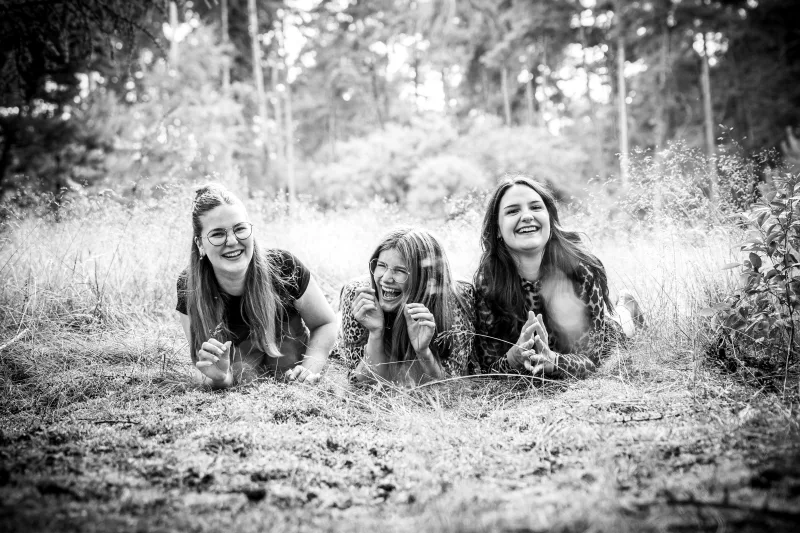 Drei lachende junge Frauen liegen im hohen Gras in einem Wald, Schwarz-Weiß-Portrait.
