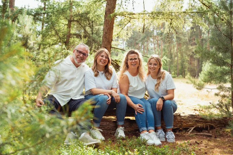 Familienfoto einer lachenden Gruppe von fünf Personen auf einem Baumstamm im Wald.