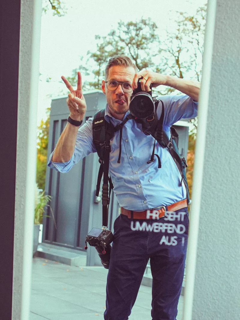 Fotograf mit Brille und professioneller Kameraausrüstung, der spielerisch ein Peace-Zeichen zeigt.