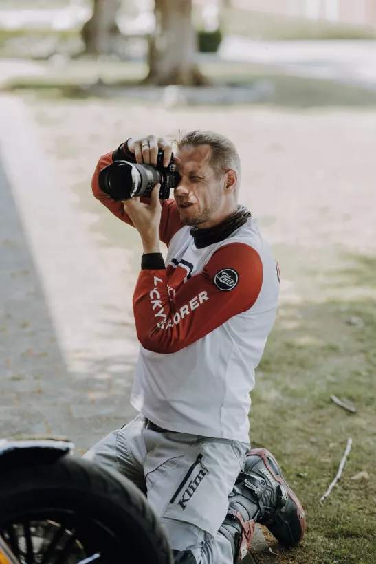 Fotograf in Motocross-Kleidung kniet auf einer Wiese und nutzt eine DSLR-Kamera