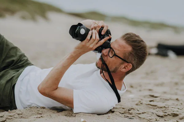 Mann blickt im Liegen am Strand durch den Sucher einer Kamera