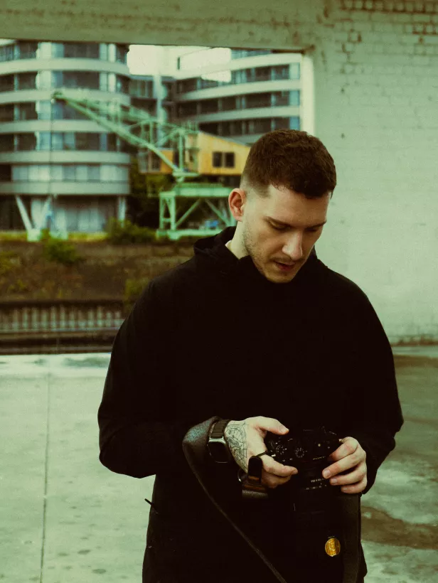 Mann in schwarzem Hoodie blickt konzentriert auf seine Kamera vor urbaner Kulisse