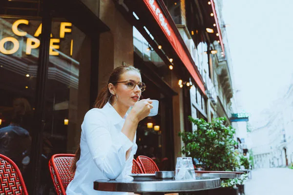 Frau mit Brille in weißer Bluse trinkt aus einer weißen Tasse an einem Außentisch eines Cafés.