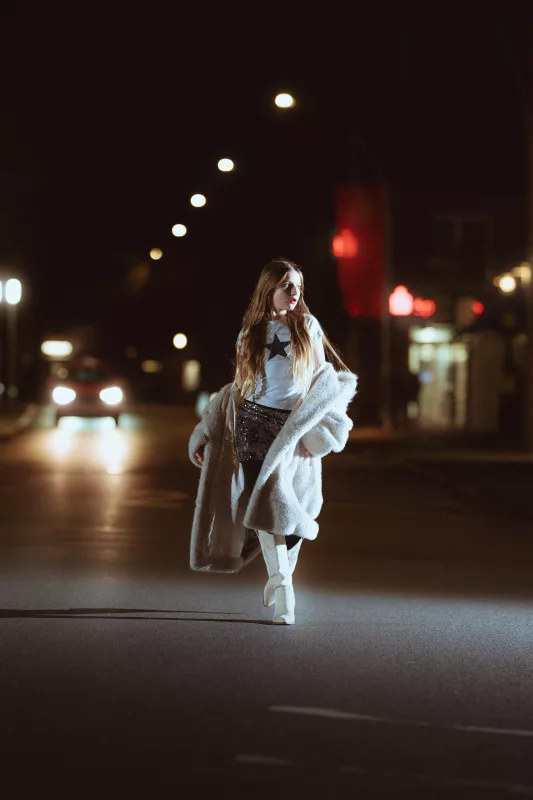 Frau in einem flauschigen weißen Mantel und weißen Stiefeln auf einer nächtlichen Straße mit Bokeh-Lichtern.