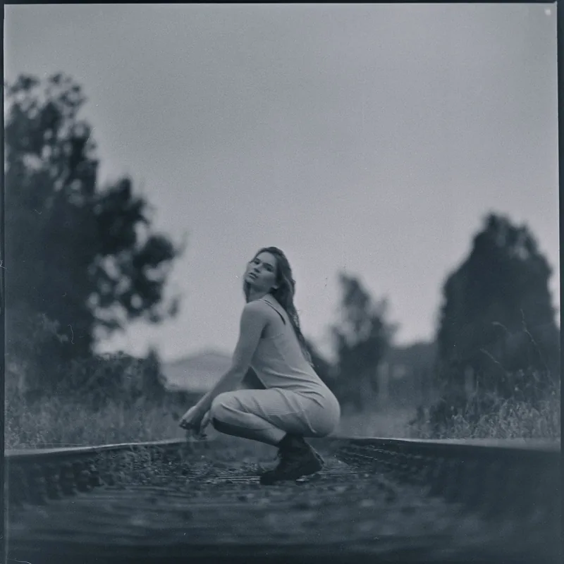 Frau in hellem Outfit hockend am Rand eines Wasserbeckens vor bewaldetem Hintergrund.