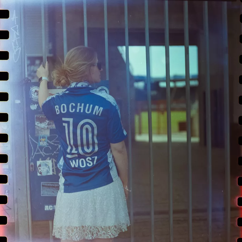 Rückansicht einer Frau im blauen Fußballtrikot und weißen Spitzenrock hinter einem Metallgitter