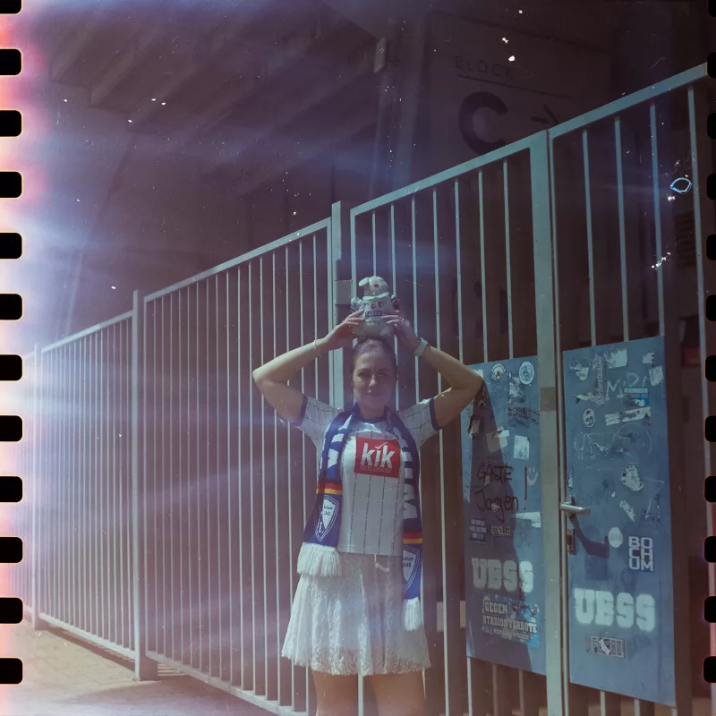 Frau in Fußballtrikot und Schal vor einem Metallzaun im Stadion