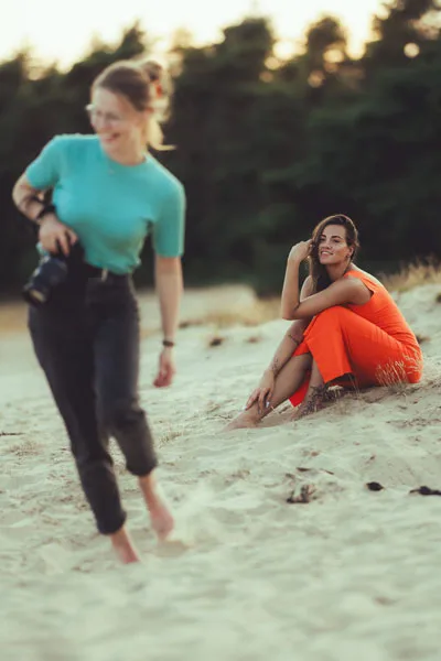 Eine Frau in einem orangefarbenen Kleid sitzt lachend auf einer Sanddüne.