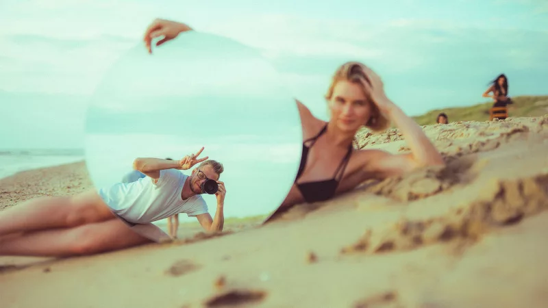 Frau im Sand mit einem runden Spiegel, der die Reflexion eines Fotografen zeigt