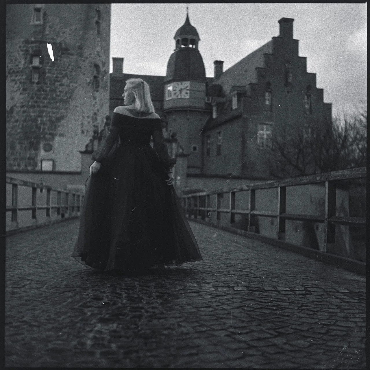 Frau in einem langen dunklen Kleid auf einer gepflasterten Brücke vor einem historischen Schlossgebäude.
