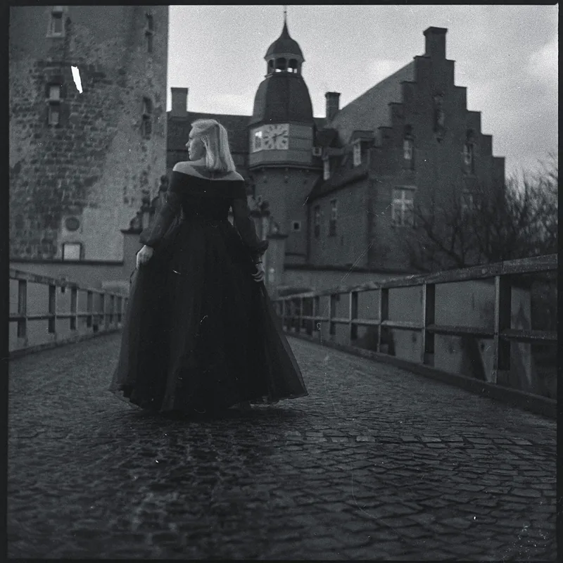 Frau in einem langen dunklen Kleid auf einer gepflasterten Brücke vor einem historischen Schlossgebäude.
