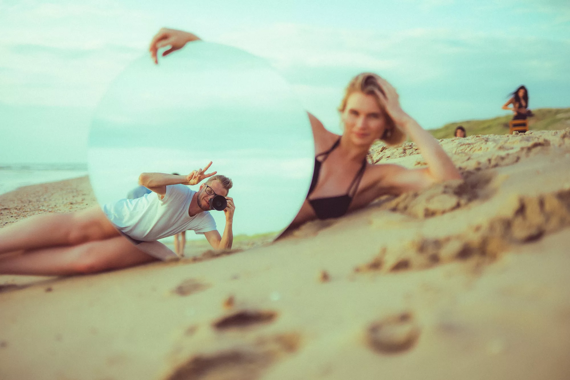 Eine Frau liegt am Strand neben einem runden Spiegel, der die Reflexion eines Fotografen zeigt.