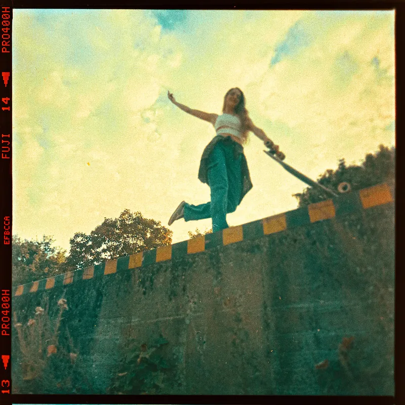 Frau mit Skateboard beim Balancieren auf einer Betonmauer vor Bäumen und Wolkenhimmel.