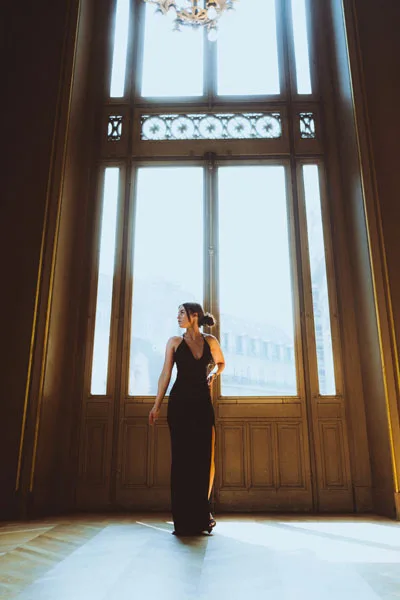 Frau in schwarzem Abendkleid vor hohen, lichtdurchfluteten Fenstern.