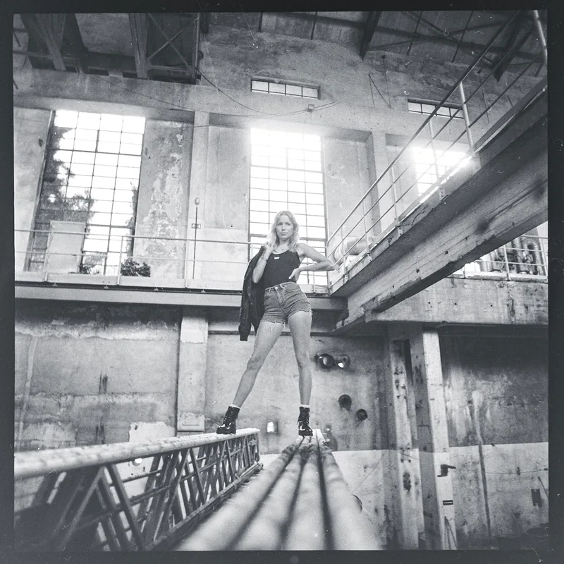 Frau in schwarzem Top und Shorts auf Metallbalken in einer Industriehalle.