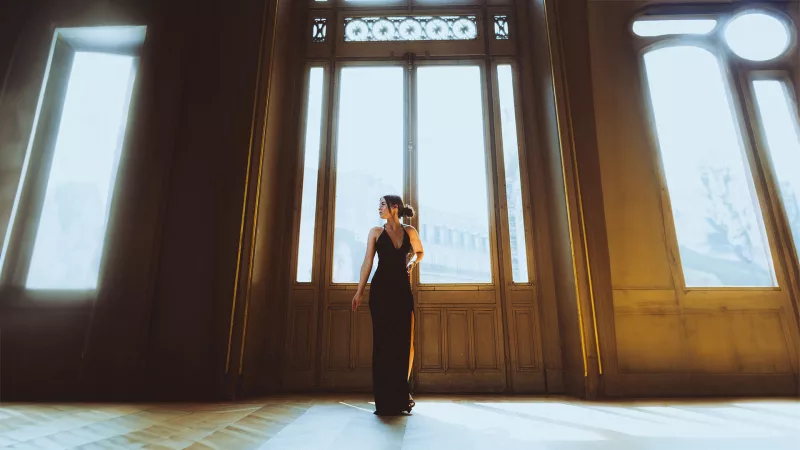 Frau in einem schwarzen Abendkleid vor hohen, lichtdurchfluteten Fenstern in Paris.