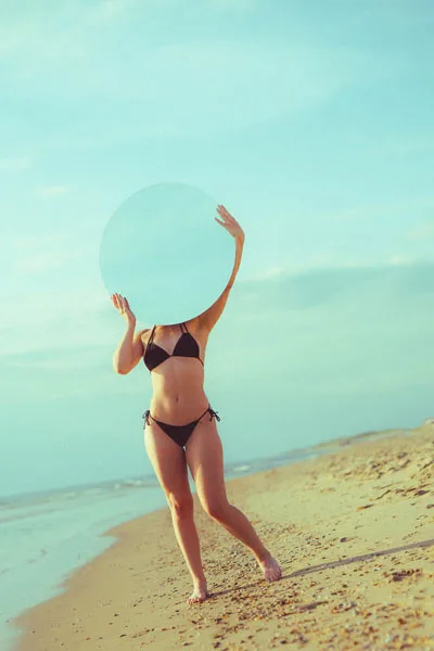 Frau im schwarzen Bikini mit einer großen runden Scheibe vor dem Gesicht am Strand.