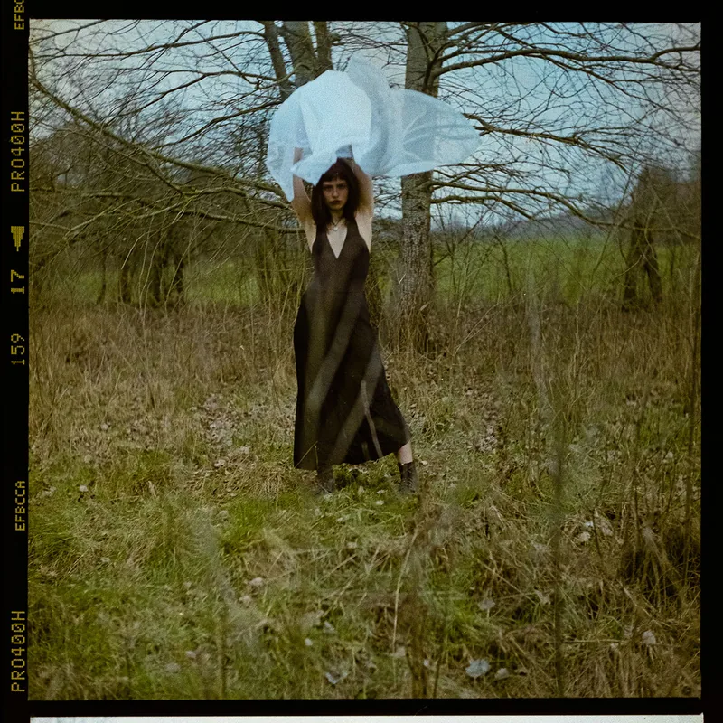 Analoges Portrait einer Frau im schwarzen Kleid mit weißem, transparentem Stoff vor kahlen Bäumen