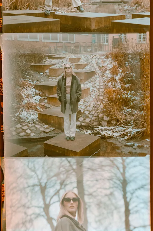 Eine Frau mit Sonnenbrille und dunkler Jacke steht auf steinernen Stufen in einer winterlichen Landschaft.