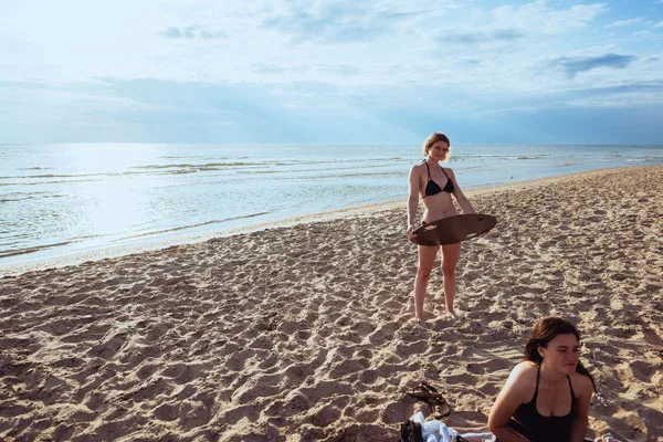 Frau mit Surfbrett am Sandstrand unter bewölktem Himmel.