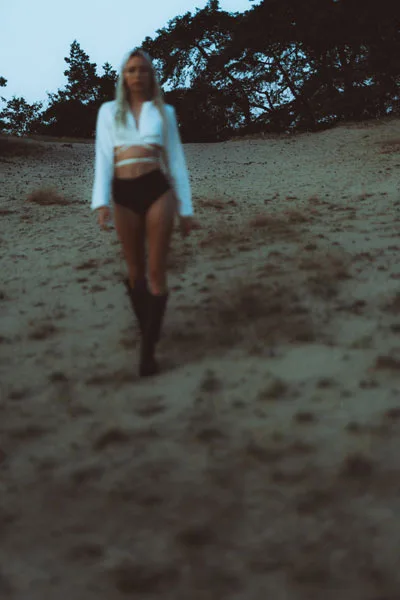 Frau in weißem Oberteil und dunkler Shorts in einer Dünenlandschaft mit Bäumen im Hintergrund.