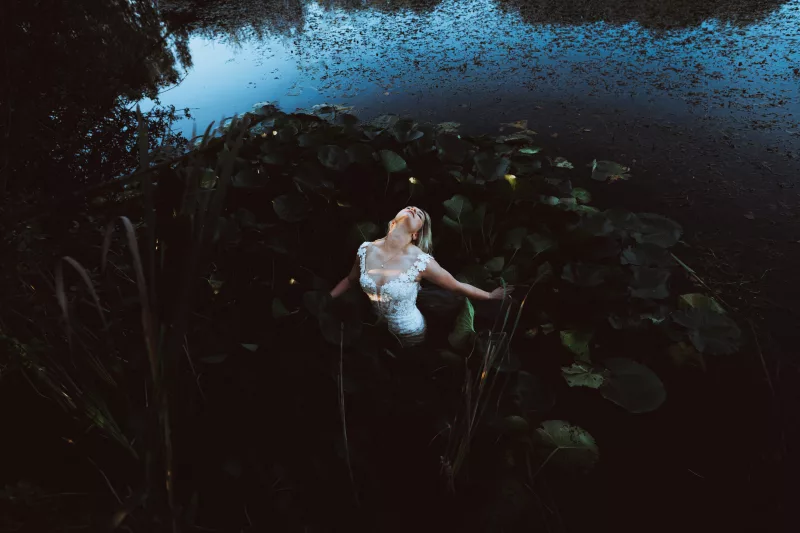 Frau in weißem Spitzenkleid inmitten von Seerosenblättern auf dunklem Wasser.