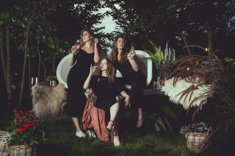 Gruppenportrait von drei Frauen in schwarzen Kleidern mit Sektgläsern vor einem weißen Sofa im nächtlichen Garten.