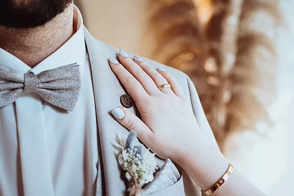 Hand mit Ehering auf dem Revers eines hellen Hochzeitsanzugs mit Fliege und Ansteckblume.