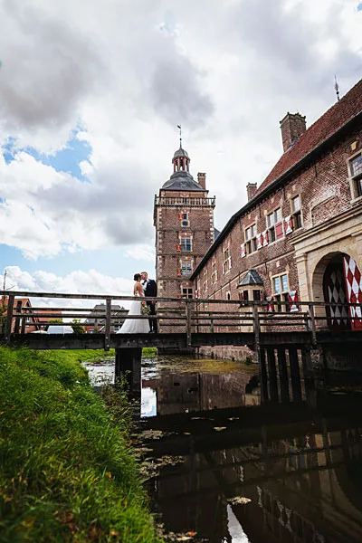 Hochzeitsfoto eines Paares auf einer Holzbrücke vor einem historischen Backsteinschloss.