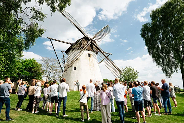 Hochzeitsgesellschaft auf einer grünen Wiese vor einer großen weißen Windmühle unter blauem Himmel.