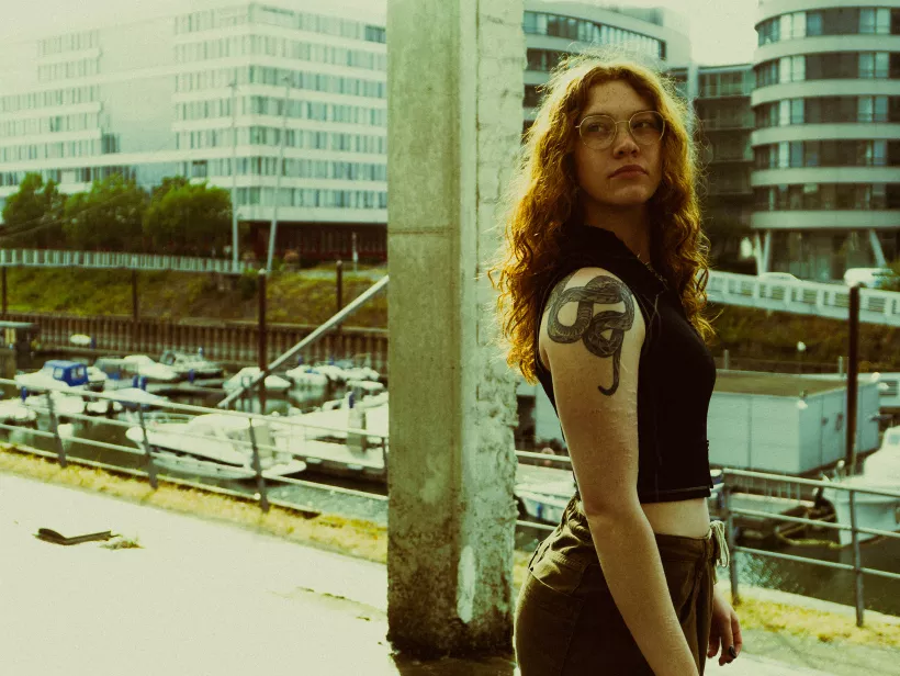 * Image Content: A portrait of a young woman with curly blonde/reddish hair, glasses, and a snake tattoo on her upper arm. She is standing