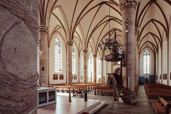 Innenraum einer gotischen Kirche mit massiven Steinsäulen, hohen Gewölben und Buntglasfenstern.