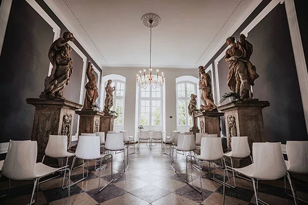 Klassische Skulpturen auf Podesten in einem prunkvollen Saal mit weißen Stühlen und Kronleuchter.