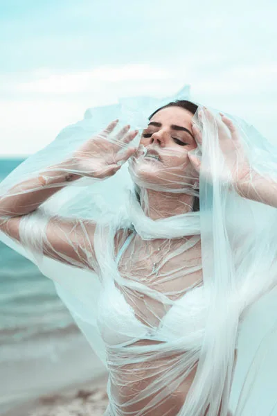 Künstlerisches Portrait einer Frau in weißem, transparentem Stoff am Strand.