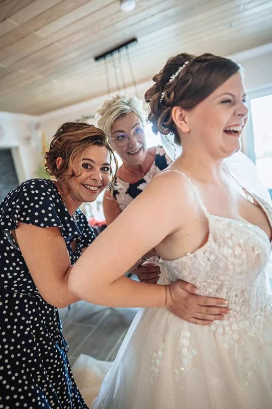 Lachende Braut in einem glitzernden Hochzeitskleid mit zwei Begleiterinnen.