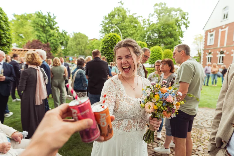 Lachende Braut im weißen Spitzenkleid mit buntem Blumenstrauß beim Anstoßen