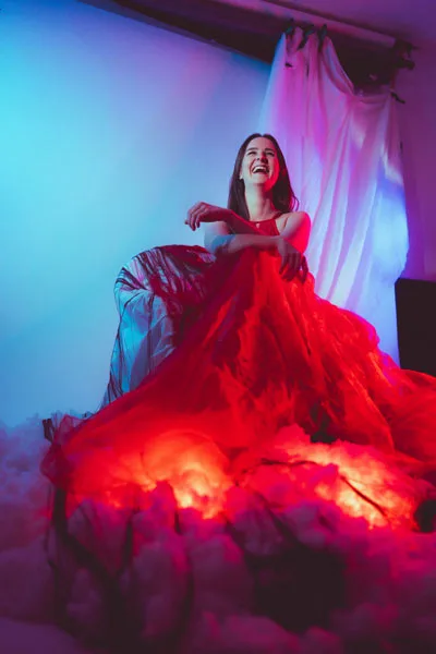 Lachende Frau in einem voluminösen roten Kleid unter dramatischem blauem und rotem Licht.