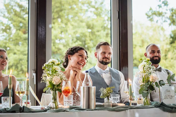 Lachende Gäste an einem festlich gedeckten Tisch mit Blumen und Kerzen.
