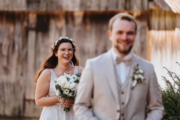 Lächelnde Braut mit Blumenkranz und weißem Bouquet in einem Hochzeitsportrait vor rustikalem Hintergrund.