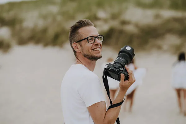 Lächelnder Mann mit Brille und Kamera in einer Dünenlandschaft