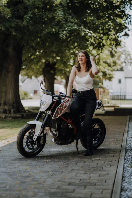 Lifestyle-Portrait einer lachenden Frau auf einem weißen Motorrad unter grünen Bäumen