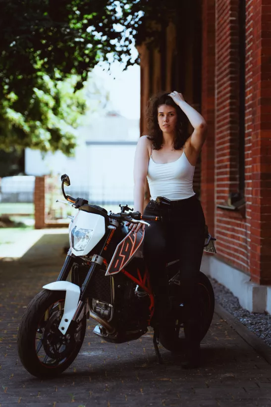 Lifestyle-Portrait einer Frau in weißem Top auf einem weißen Motorrad vor einer Backsteinwand