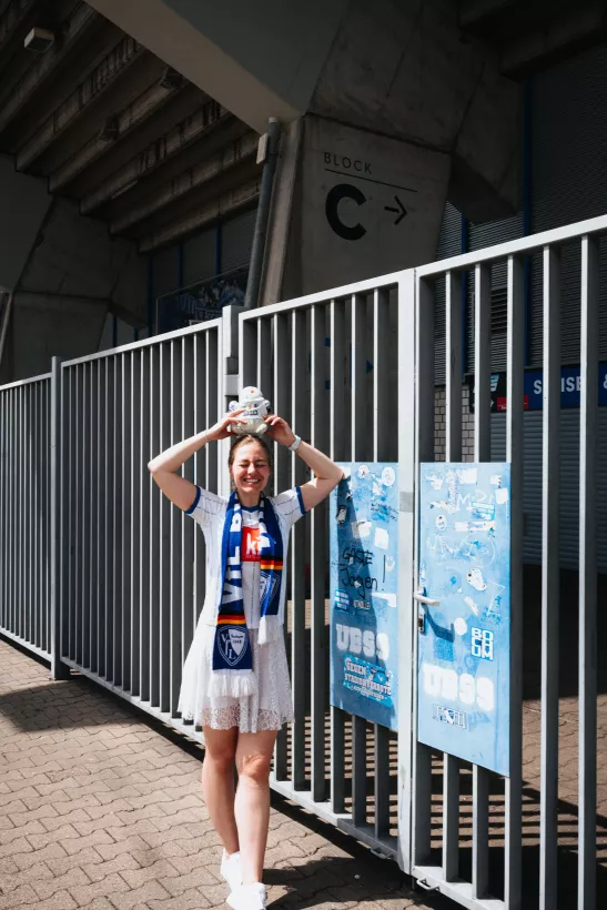 Outdoor-Portrait einer lächelnden Frau mit Fan-Schal vor einem Metallgitter am Stadioneingang