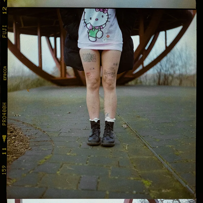Person in einem Hello-Kitty-Shirt und schwarzen Boots mit sichtbaren Tätowierungen an den Oberschenkeln unter einer Holzkonstruktion.