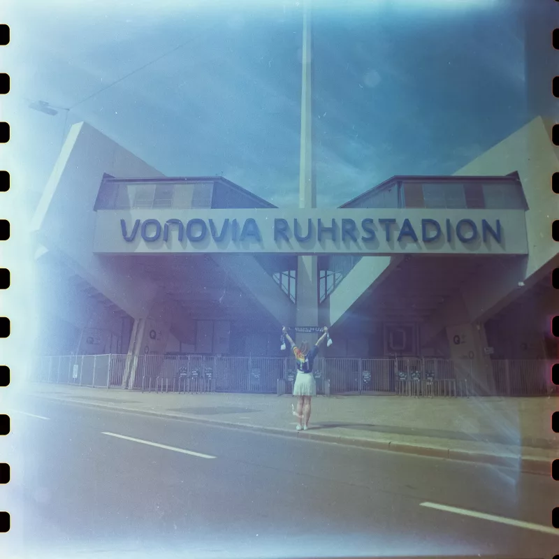 Person mit erhobenen Armen vor dem Vonovia Ruhrstadion in analoger Optik